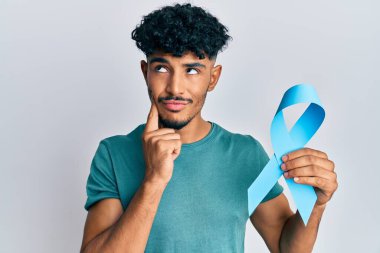 Genç Arap yakışıklı adam mavi kurdele tutuyor ciddi bir yüzü var eli çenesinde soru sormayı düşünüyor, kafa karıştırıcı bir fikir. 