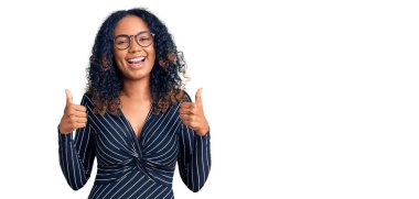 Genç Afro-Amerikalı kadın günlük kıyafetler ve gözlük takıyor. Başarı işareti el hareketleriyle olumlu jest yapıyor. Gülümsüyor ve mutlu. Neşeli ifade ve kazanan jest. 