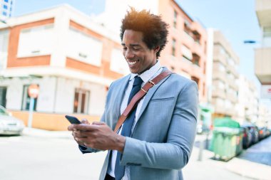 Genç Afrikalı Amerikalı iş adamı mutlu bir şekilde gülümsüyor. Yüzünde gülümsemeyle şehrin caddesinde akıllı telefon kullanarak dikiliyorsun..