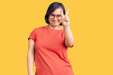 Down sendromlu esmer kadın sıradan kıyafetler giyiyor ve gözlük takıyor. Kameraya gülümsüyor ve zafer işareti yapıyor. İki numara.. 