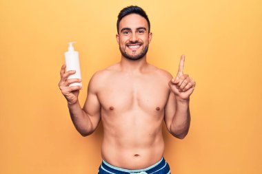 Tatilde sakallı yakışıklı bir adam elinde mayo, elinde güneş kremi şişesi bir fikirle gülümsüyor ya da soru işareti yapıyor.