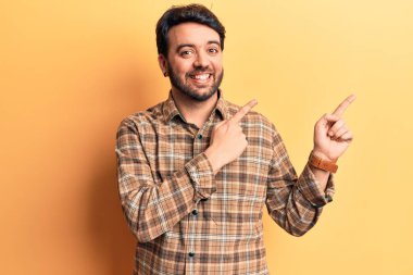 Gündelik kıyafetler giyen genç İspanyol adam gülümseyip kameraya bakıyor ve iki eliyle ve parmaklarıyla kenarı işaret ediyor.. 