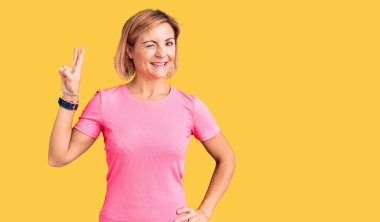 Spor kıyafetleri giyen, gülümseyen, kameraya göz kırpan ve parmaklarıyla zafer işareti yapan genç sarışın kadın. İki numara.. 