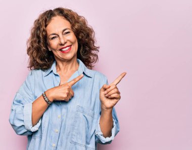 Orta yaşlı güzel bir kadın pembe arka planda dikilip gülümseyen ve kameraya bakıp iki elini ve parmaklarını yana doğru gösteren sıradan bir kot gömlek giyiyor..