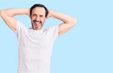 Orta yaşlı yakışıklı bir adam günlük tişörtler giyiyor rahatlıyor ve esniyor, kolları ve elleri başının arkasında mutlu bir şekilde gülümsüyor. 