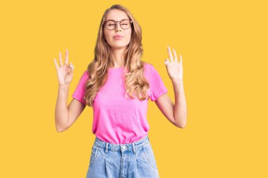 Sarı saçlı, sıradan kıyafetler giyen, gözlüklü, gözleri kapalı gülümseyen, meditasyon hareketi yapan genç, beyaz bir kadın. yoga konsepti. 