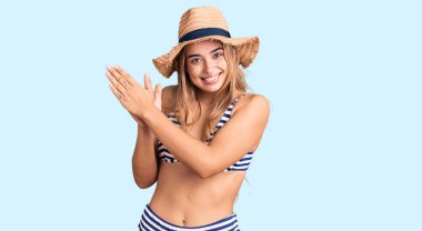 Bikini giyen, şapka takan, alkışlayan mutlu ve neşeli gülümseyen güzel sarışın bir kadın. 