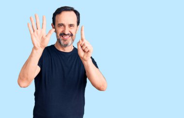 Orta yaşlı yakışıklı bir adam gündelik tişört giyer ve altı numaralı parmaklarıyla işaret ederken kendinden emin ve mutlu bir şekilde gülümser.. 