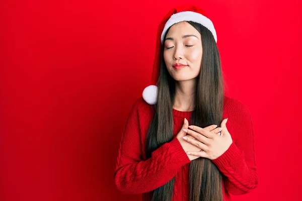 若い中国の女性の胸に手で笑みを浮かべてクリスマスの帽子を着て 目の顔に感謝のジェスチャーで閉じます 健康の概念 — ストック写真