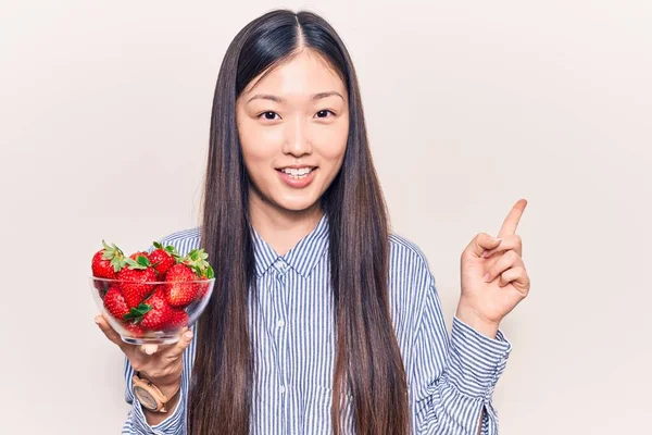 年轻美丽的中国女人拿着一碗草莓微笑着 手指手画脚地指向旁边 — 图库照片