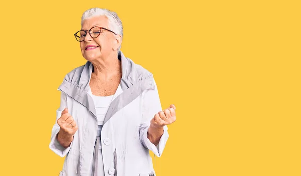 Senior beautiful woman with blue eyes and grey hair wearing casual clothes and glasses very happy and excited doing winner gesture with arms raised, smiling and screaming for success. celebration concept.