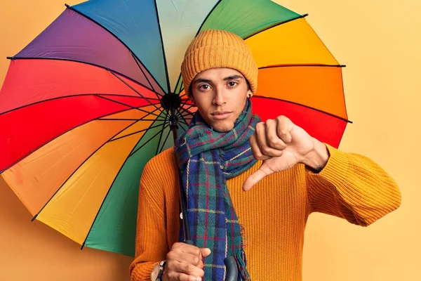 年轻的非洲裔美国人拿着五颜六色的雨伞 满脸怒容 消极的迹象表明他不喜欢大拇指朝下 拒绝的概念 — 图库照片