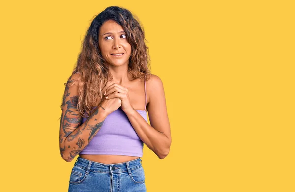 Young hispanic woman with tattoo wearing casual clothes laughing nervous and excited with hands on chin looking to the side