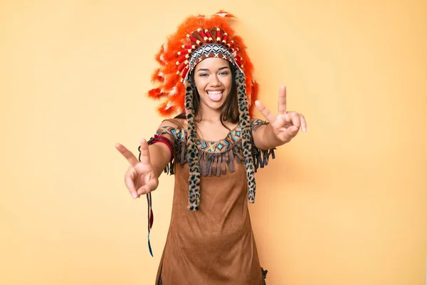 Young beautiful latin girl wearing indian costume smiling with tongue out showing fingers of both hands doing victory sign. number two.
