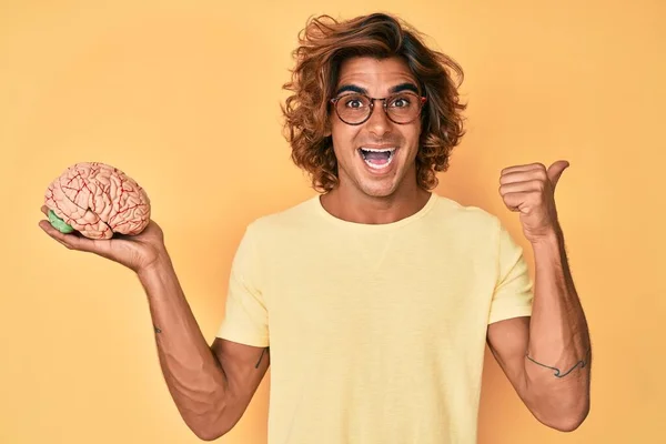 Jovem Hispânico Segurando Cérebro Apontando Polegar Para Lado Sorrindo Feliz — Fotografia de Stock