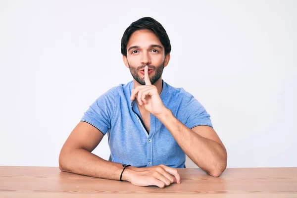 テーブルの上に座るカジュアルな服を着たハンサムなヒスパニック系の男が唇に指で静かにするように求めている 沈黙と秘密の概念 — ストック写真