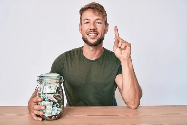 Joven Hombre Caucásico Sosteniendo Frasco Con Ahorros Sentado Mesa Sonriendo —  Fotos de Stock