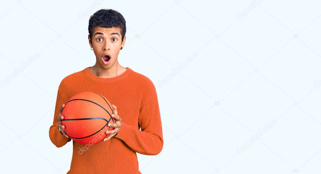 Young african amercian man holding basketball ball scared and amazed with open mouth for surprise, disbelief face 