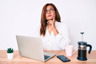 Orta yaşlı esmer, İspanyol iş kadını ofiste çalışıyor ciddi bir yüz ifadesi var eli çenesinde soru soran, kafa karıştırıcı bir fikir hakkında düşünceli. 