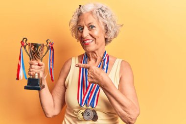 Kıdemli gri saçlı kadın elinde şampiyonluk kupası, madalyalar takıyor mutlu bir şekilde el ve parmak işareti yapıyor. 