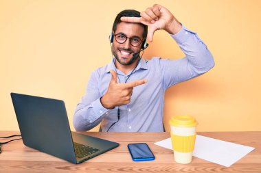 Masada oturan İspanyol genç adam çağrı merkezinde operatör kulaklığı takıyor gülümsüyor ve elleri ve parmakları gülen yüzüyle çerçeve yapıyor. yaratıcılık ve fotoğrafçılık kavramı. 