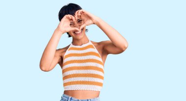 Kısa saçlı genç esmer kadın gündelik giysiler giyiyor kalp şeklinde el ve parmaklarıyla tabelanın içinden bakıp gülümsüyor. 