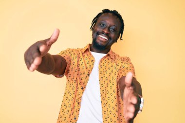 Saçları örgülü genç Afro-Amerikan bir adam. Sıradan kıyafetler giyip kameraya bakıyor. Sarılmak için kollarını açmış gülümsüyor. Mutluluğu kucaklayan neşeli bir ifade.. 