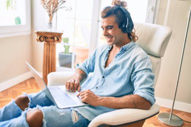 Evde dizüstü bilgisayar ve kulaklık kullanarak mutlu gülümseyen genç İspanyol adam.