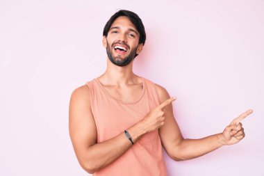 Yakışıklı, sıradan kıyafetler giyen, gülümseyen ve kameraya bakan, iki elini ve parmağını yana doğru gösteren İspanyol bir adam.. 