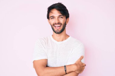 Yakışıklı, beyaz tişörtlü mutlu yüzlü, kollarını kavuşturup kameraya bakan bir İspanyol. pozitif kişi. 