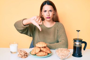Genç esmer kadın masada oturmuş kızgın yüzüyle kahvaltı yapıyor. Negatif işaret baş parmakları aşağıda, reddedilme konsepti ise... 