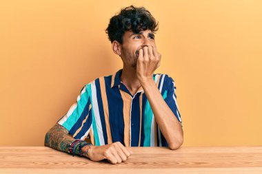 Masada oturan Latin asıllı genç adam gergin ve gergin görünüyor. Ağzı tırnak yiyen ellerle. anksiyete. 