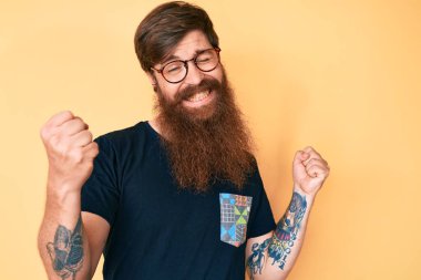Uzun sakallı, sıradan kıyafetler ve gözlük takan yakışıklı kızıl saçlı, çok mutlu ve heyecanlı bir adam. Kolları havada, gülümseyen ve başarı için çığlık atan, kazanan bir jest yapıyor. kutlama konsepti. 