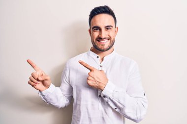 Sakallı, sıradan tişörtlü genç bir adam beyaz arka planda dikiliyor gülümsüyor ve kameraya bakıyor iki eliyle ve parmaklarıyla yan tarafa bakıyor..