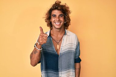 Yaz stili giyinmiş genç İspanyol adam mutlu ve pozitif gülümsüyor, başparmağıyla mükemmel ve onay işareti yapıyor. 