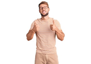 Gündelik kıyafetler ve gözlük takan beyaz bir genç. Başarı işareti ile el hareketi yapıyor. Gülümsüyor ve mutlu. Neşeli ifade ve kazanan jest. 