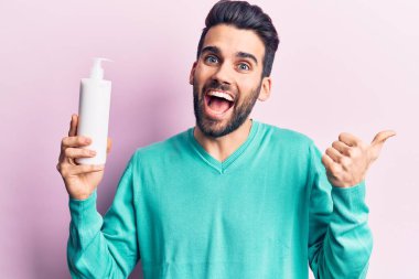Sakallı, sakallı, elinde krema şişesi olan genç ve yakışıklı bir adam baş parmağını kaldırmış, ağzı açık gülümsüyor. 