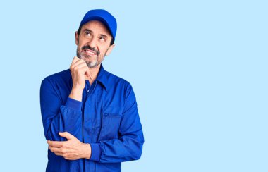 Orta yaşlı yakışıklı, elleri çenesinde tamirci üniforması giyen soru soran, düşünceli bir adam. Gülümseyen ve düşünceli bir yüz. şüphe kavramı. 