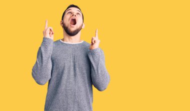 Gündelik süveter giyen genç yakışıklı adam şaşırmış ve şaşırmış bir şekilde yukarı bakıyor ve parmaklarıyla işaret ediyor ve kollarını kaldırıyor.. 