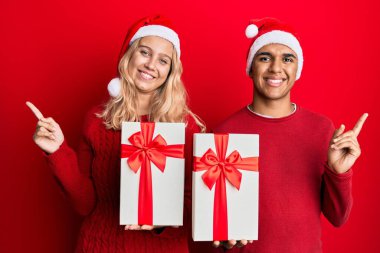 Irklar arası genç bir çift Noel şapkası takıyor ve bir hediye tutuyorlar mutlu bir şekilde gülümsüyorlar. 