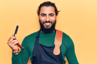 Önlük giyen genç Arap adam berber jileti tutuyor. Olumlu ve mutlu görünüyor. Kendine güvenen bir gülümsemeyle dişlerini gösteriyor. 