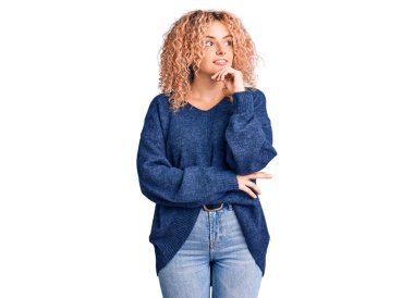 Kıvırcık saçlı, sıradan bir kazak giyen, eli çenesinde soru soran genç sarışın kadın, düşünceli bir ifade. Gülümseyen ve düşünceli bir yüz. şüphe kavramı. 