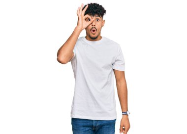 Gündelik beyaz tişört giyen genç Arap adam şaşırmış yüz ifadesiyle şaşırmış bir şekilde hareket ediyor ve parmaklarının arasından bakıyor. İnançsız ifade. 