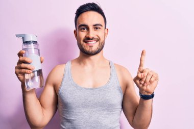 Sakallı, kolsuz tişörtlü genç yakışıklı bir sporcu bir fikirle ya da soruyla gülümseyen bir şişe su içiyor.
