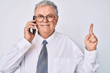 Kıdemli gri saçlı adam iş kıyafetleri giymiş akıllı telefonda konuşuyor bir fikir veya soruyla gülümsüyor ve mutlu bir yüzle işaret ediyor. 