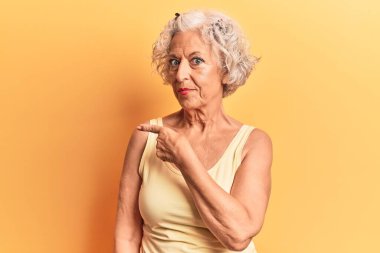 Yaşlı, gri saçlı, günlük kıyafetler giyen, el parmaklarıyla işaret eden, ciddi ve sakin bir yüz ifadesi olan bir kadın. 