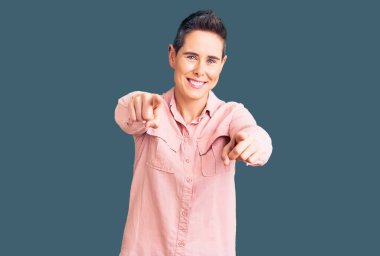 Kısa saçlı, günlük kıyafetler giyen genç bir kadın seni ve kamerayı parmaklarıyla göstererek mutlu ve mutlu bir şekilde gülümsüyor. 