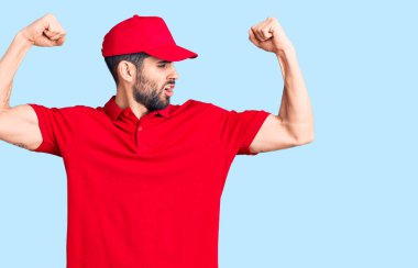 Sakallı, teslimat üniforması giyen genç yakışıklı adam gururla gülümseyen kol kaslarını gösteriyor. fitness konsepti. 