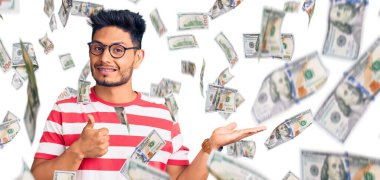 Yakışıklı Latin Amerikalı genç adam, günlük kıyafetler ve gözlük takıyor, avuç içi gösteriyor ve baş parmaklarıyla hareket ediyor, mutlu ve neşeli gülümsüyor.