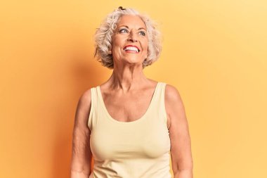 Yaşlı, gri saçlı, günlük kıyafetler giyen, yüzünde gülümsemeyle yan yana bakan bir kadın, doğal bir ifade. Kendinden emin gülen. 